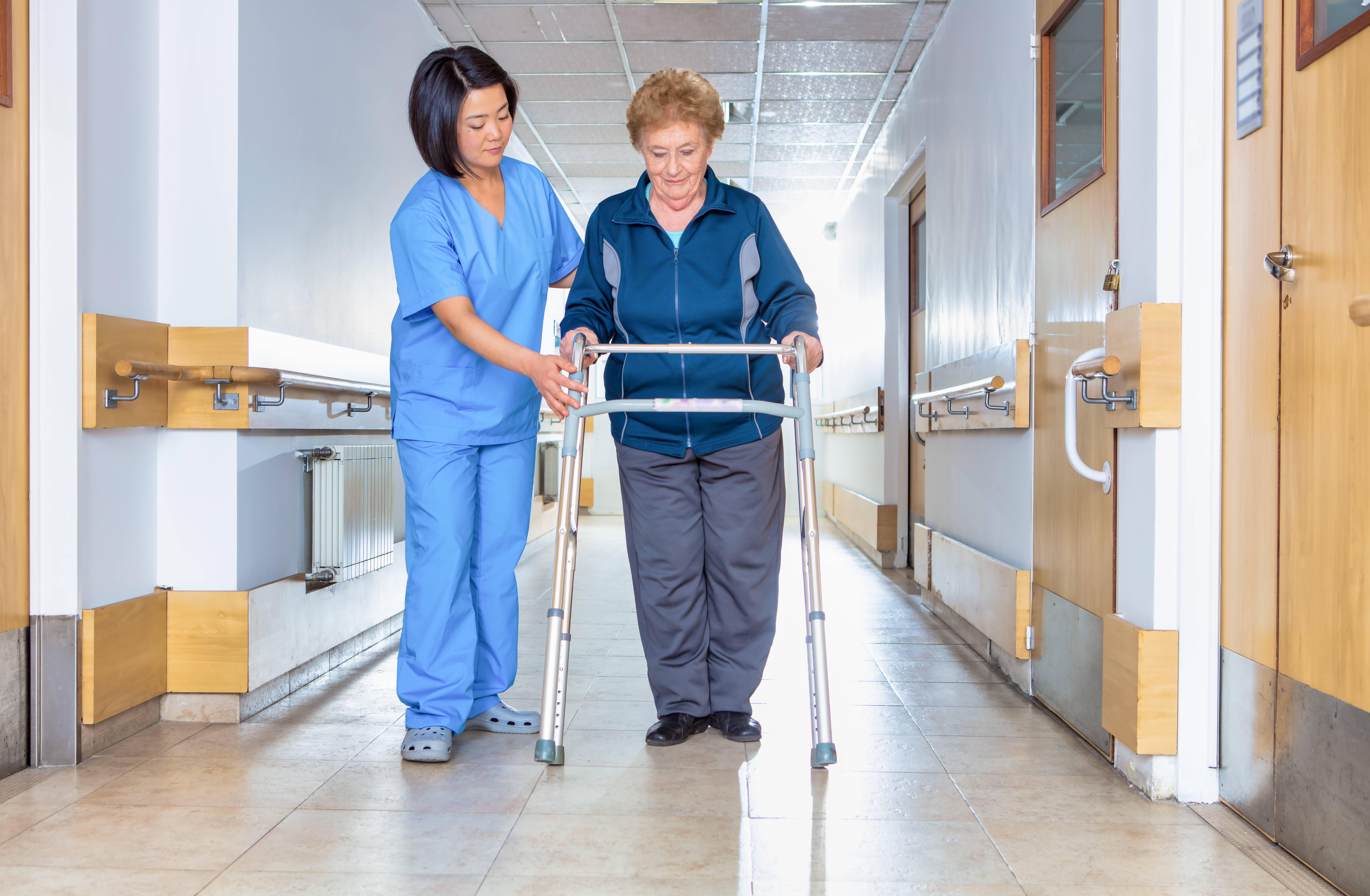 A female healthcare professional dressed in hospital attire assisting an elderly female patient with a walker in the hallway of a hospital.