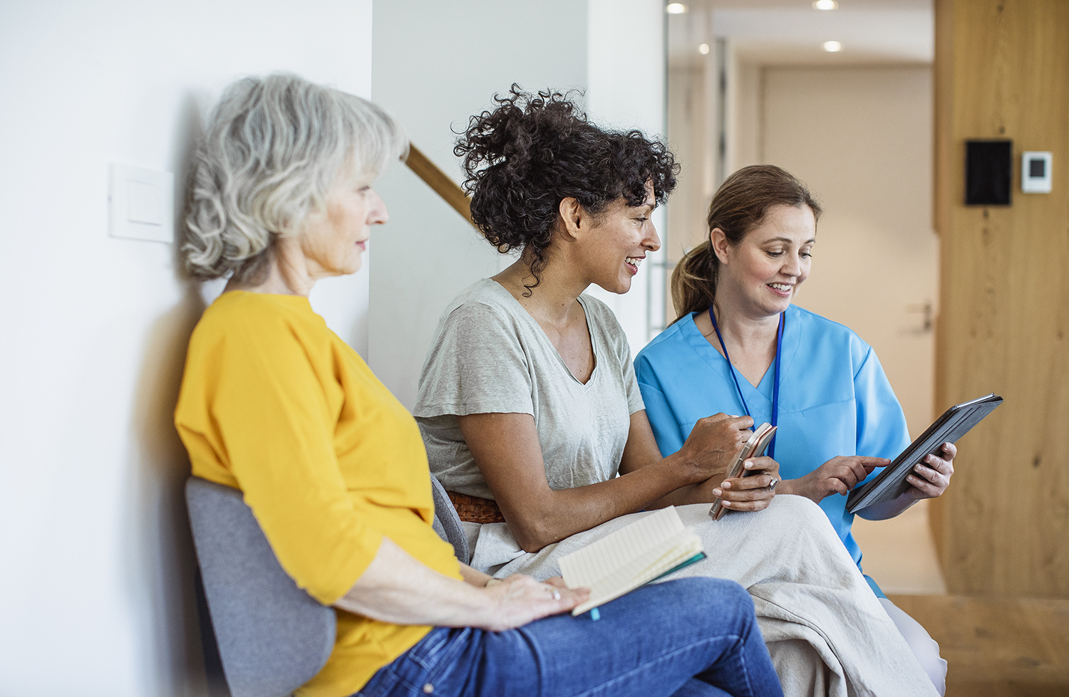 Une prestataire de soins de santé regarde une tablette avec sa patiente tandis qu’une autre patiente est assise à côté d’elles et lit un livre.