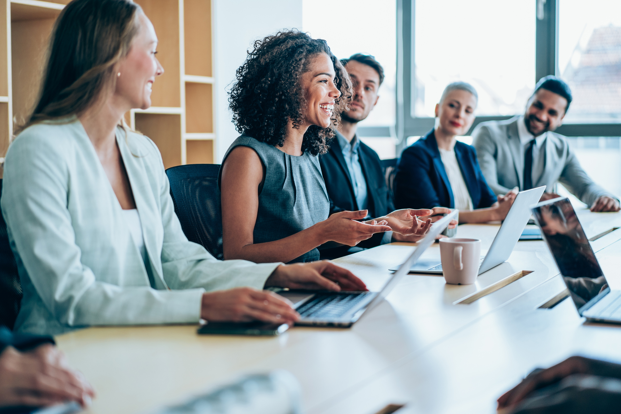 A group of five board members, two male and three female sitting at a table with laptops all focused on one female who is speaking.