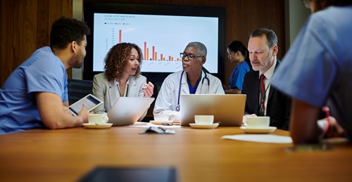 A group of healthcare professionals and administrators gathered around a laptop in a meeting room.