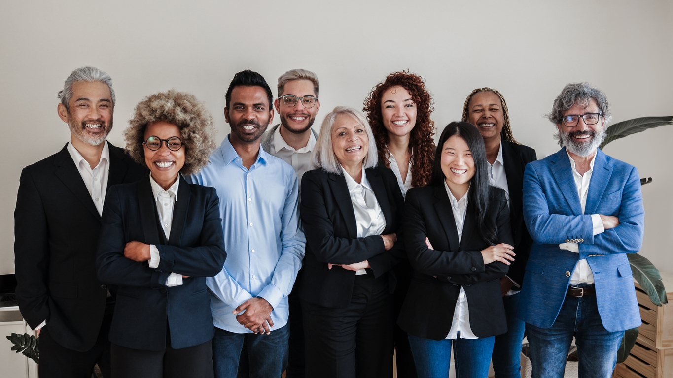 Un groupe diversifié composé de neuf personnes, dont quatre hommes et cinq femmes, se tiennent debout et sourient à la caméra.