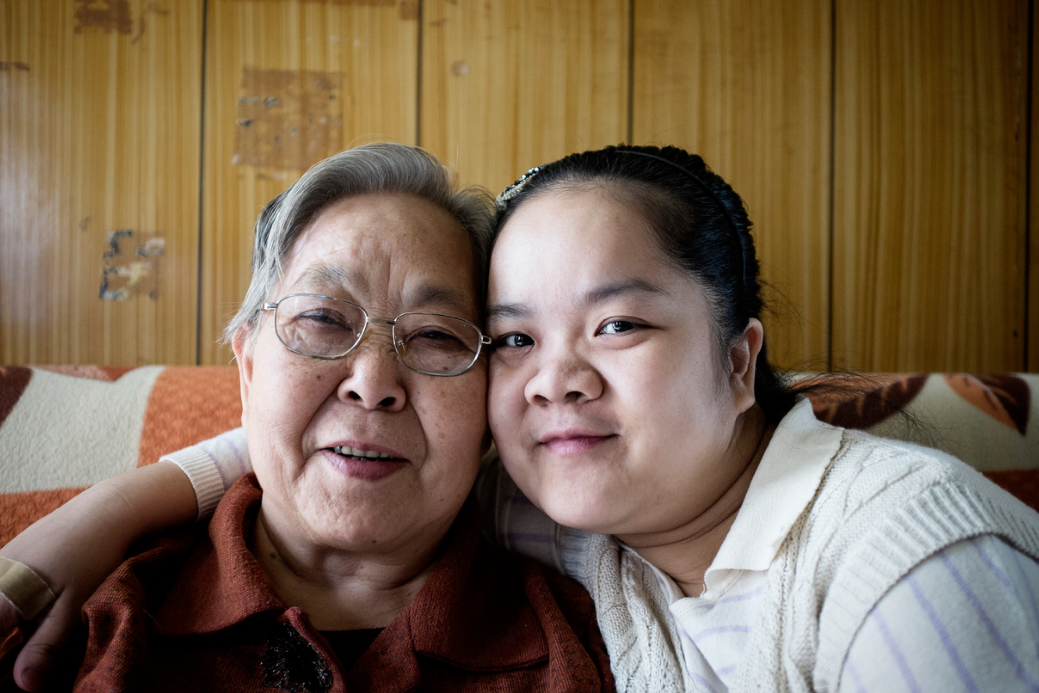 A younger person sits beside an older person on a couch with their arm around them.