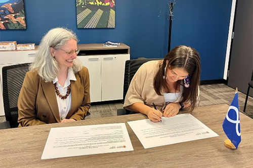 Deux personnes assises à une table de conférence, signant une entente de partenariat. L’une d’elles sourit en regardant l’autre signer. Un petit drapeau de la nation métisse est posé sur la table devant les documents.