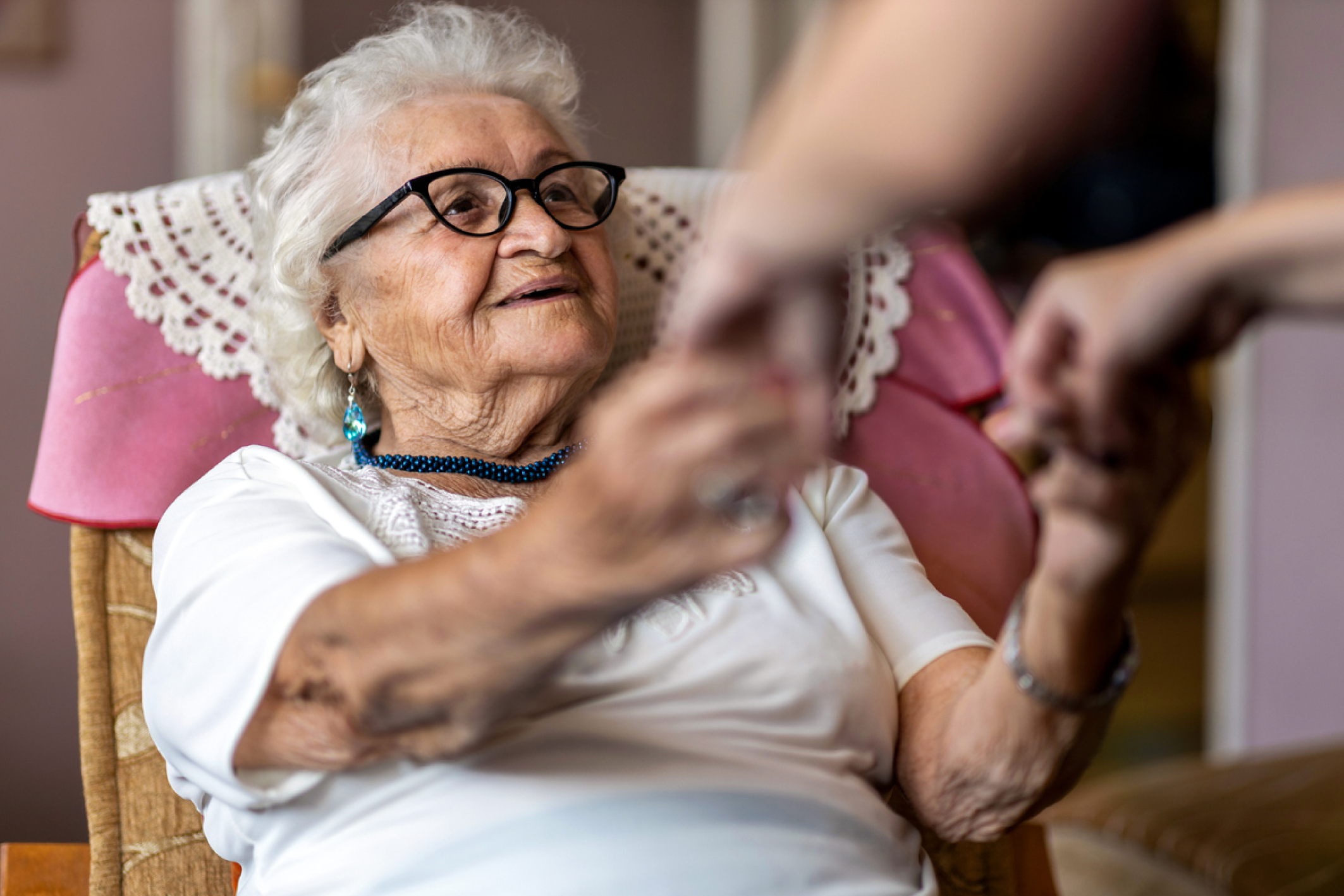 A home care worker helps an older adult stand up from an armchair.