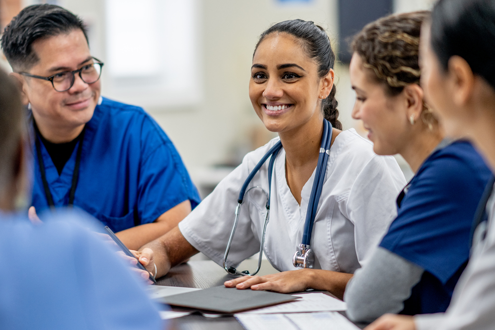 A small medical staff team sit around a boardroom table as they meet to discuss and collaborate. They are each dressed professionally and are focused on the meeting.