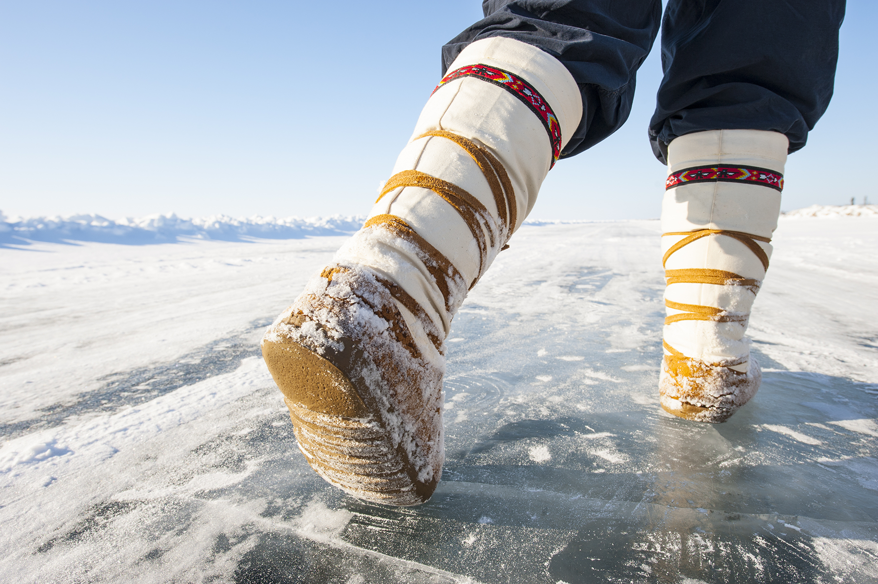 A closeup of a person’s Mukluks while they’re walking on an ice road.