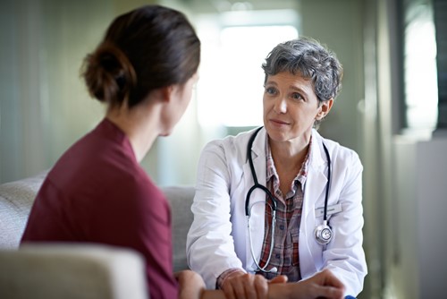 A female healthcare professional sitting closely beside a female patient having a conversation in a hospital waiting room.